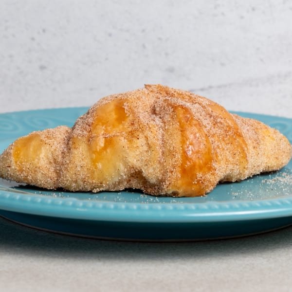 A golden-brown croissant, generously coated with cinnamon sugar, rests on a bright blue plate.